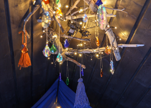 Dekorativ ljuskrona av drivved med hängande glaspärlor och små ljusslingor i varma färger mot ett mörkt träinnertak.