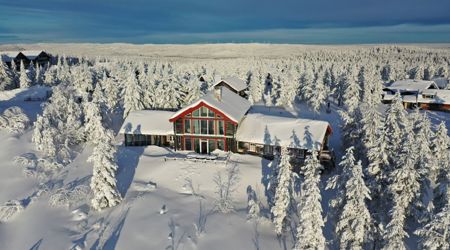Luftfoto af en stor fjeldhytte omgivet af snedækkede træer og et vidtstrakt vinterlandskab under klar himmel.