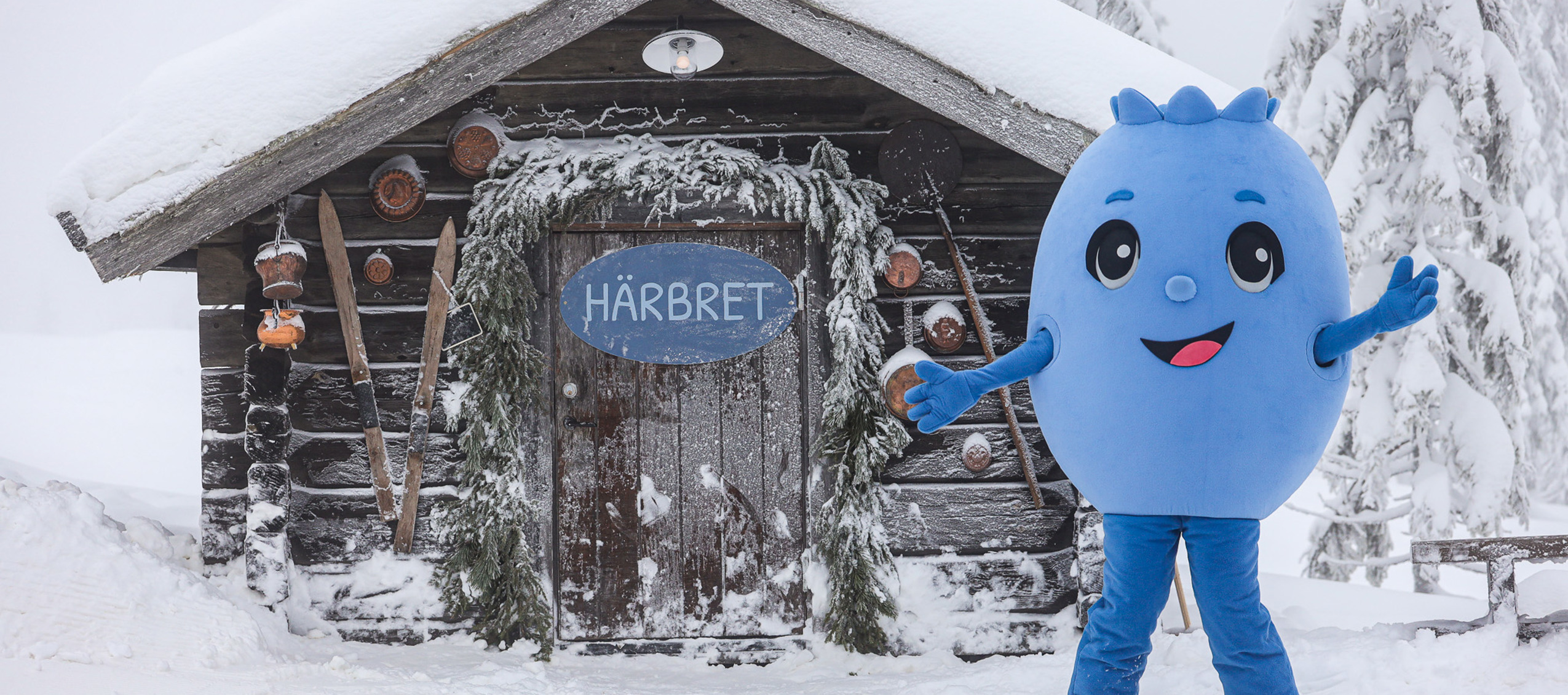 Blå maskot på skidor framför en snötäckt trästuga med skylten Härbret.