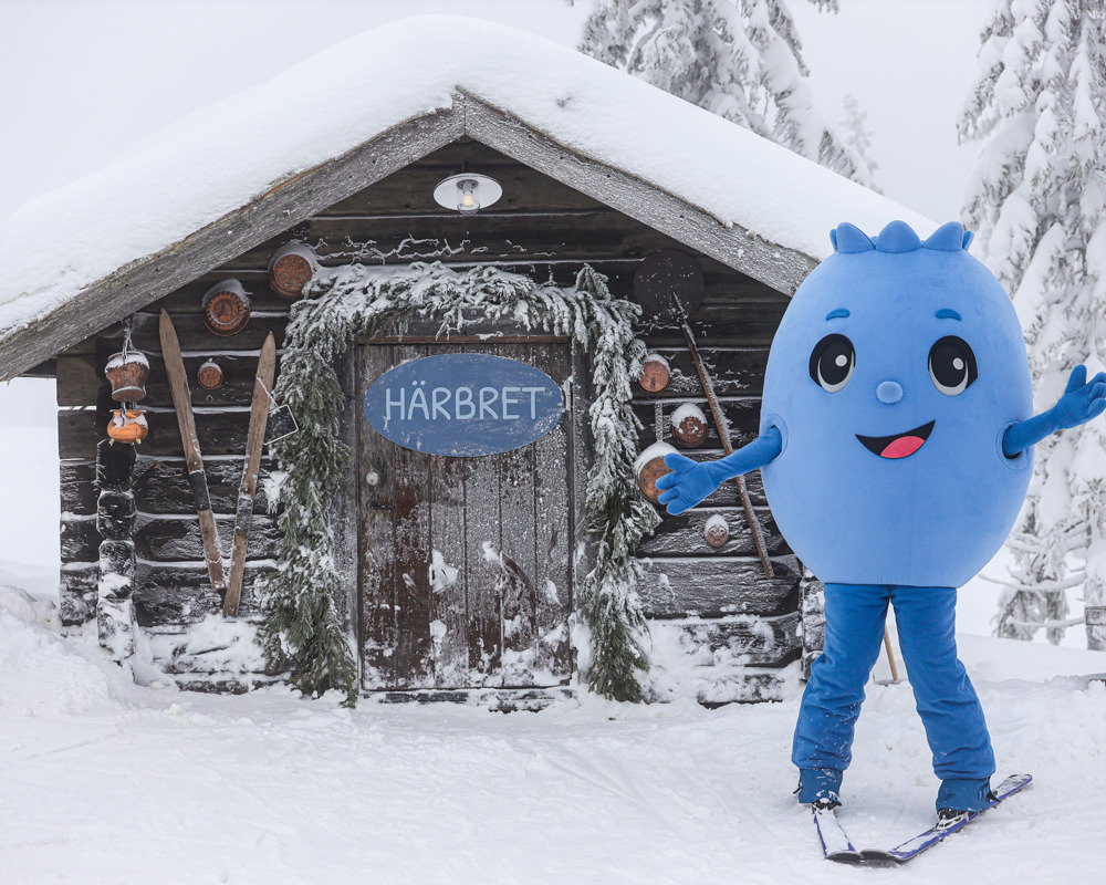 Blå maskot på skidor framför en snötäckt trästuga med skylten Härbret.