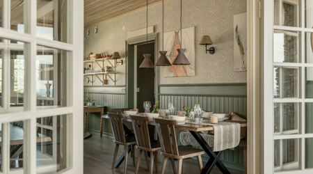 Spisestue med dækket træbord, grønne panelvægge, hængende lamper i brune nuancer og dobbeltdøre åbne til et tilstødende rum.