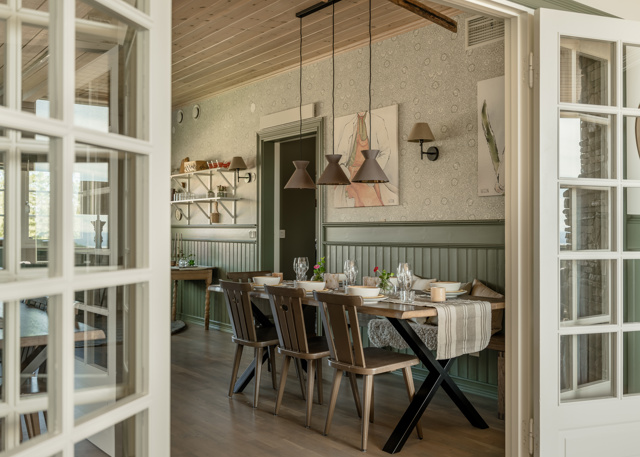 Matrum med dukat trädgårdsbord, gröna panelväggar, hängande lampskärmar i bruna toner och öppna dubbeldörrar mot ett angränsande rum.