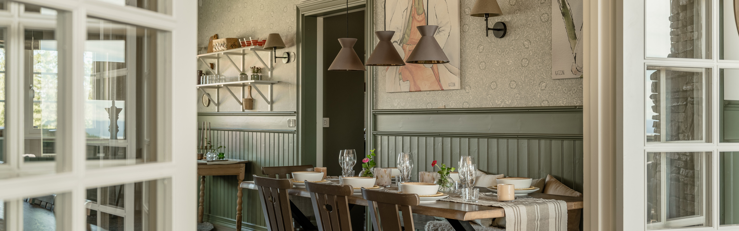 Matrum med dukat trädgårdsbord, gröna panelväggar, hängande lampskärmar i bruna toner och öppna dubbeldörrar mot ett angränsande rum.