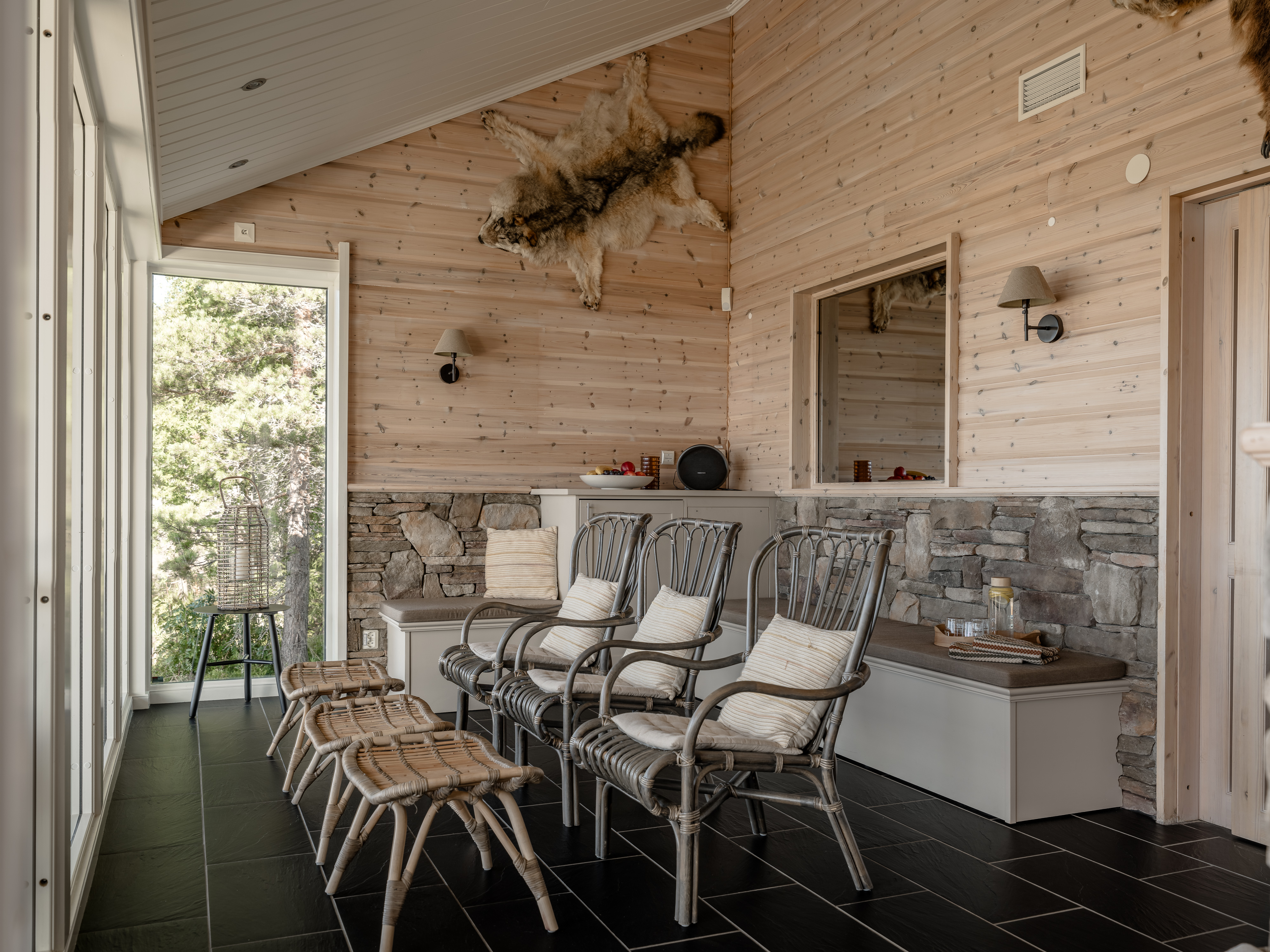 Ljus relaxavdelning med stora fönster, rottingstolar med kuddar, små pallar och bänkar längs väggar av trä och natursten. En fäll hänger på väggen och ett fat med glas och dryck står på en bänk.