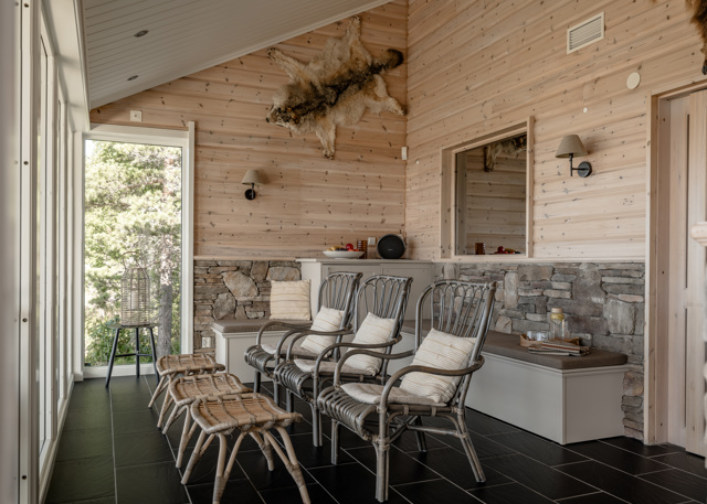 Ljus relaxavdelning med stora fönster, rottingstolar med kuddar, små pallar och bänkar längs väggar av trä och natursten. En fäll hänger på väggen och ett fat med glas och dryck står på en bänk.