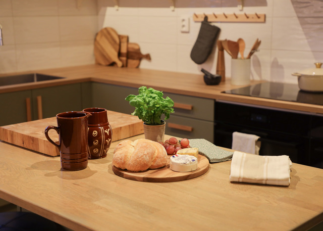 Køkkenbord med brød, ost, krydderurter og krus i et hyggeligt køkken med trædetaljer.