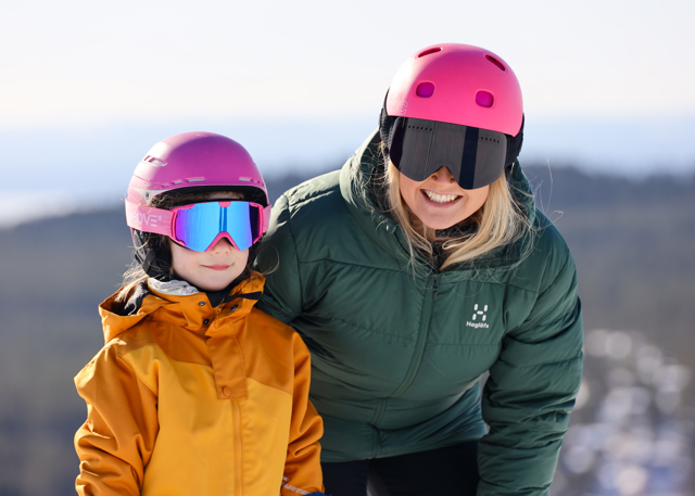 Smilende voksen og barn i lyserøde hjelme poserer sammen i skibakken med udsigt over dalen.