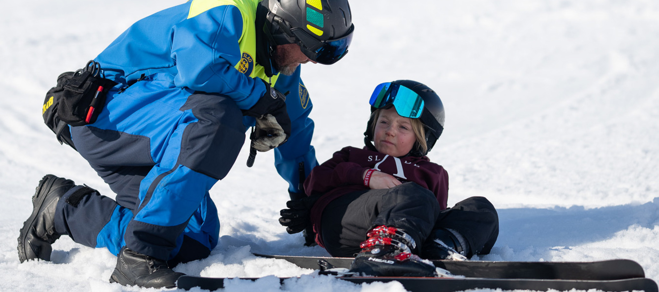 En skidlärare i blå jacka hjälper ett barn som sitter i snön med skidor på fötterna.