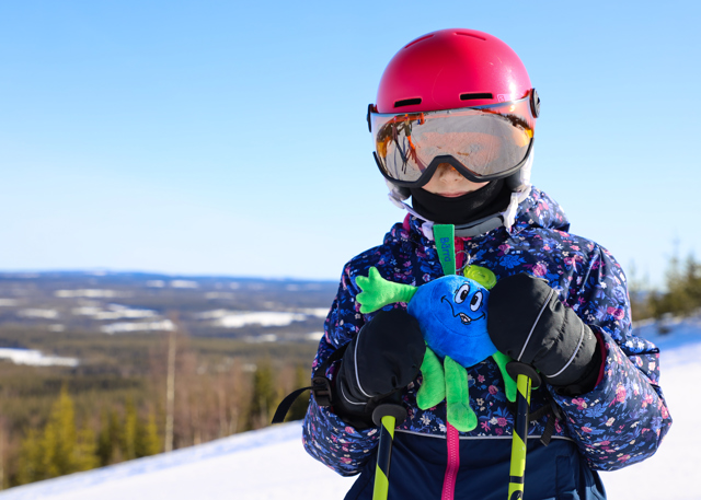 Ett barn med röd hjälm och blommig skidjacka håller i ett mjukisdjur föreställande blåbäret Bärra, med skog och fjäll i bakgrunden.