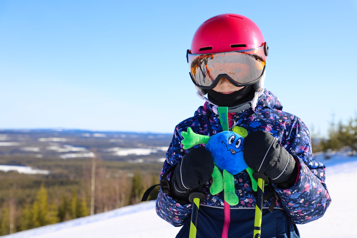 Ett barn med röd hjälm och blommig skidjacka håller i ett mjukisdjur föreställande blåbäret Bärra, med skog och fjäll i bakgrunden.