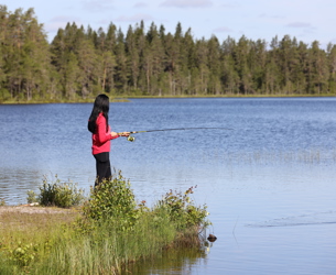 Kvinna i röd tröja fiskar vid en stilla sjö omgiven av skog.
