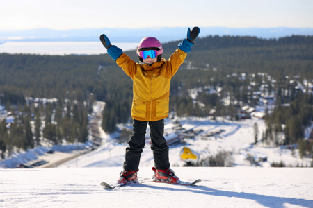 Child in colorful ski clothes stands with arms raised at the top of a ski slope with snowy landscape in the background.