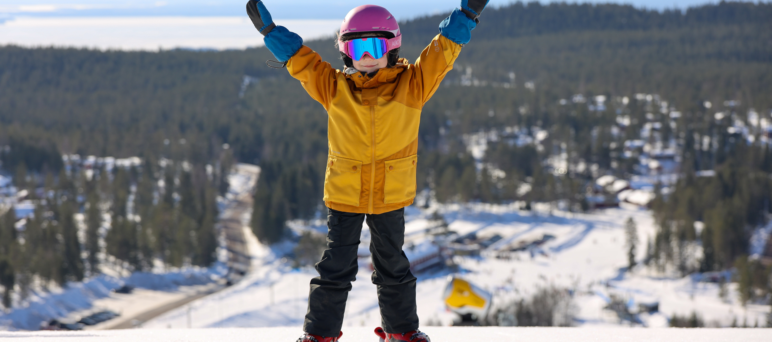 Child in colorful ski clothes stands with arms raised at the top of a ski slope with snowy landscape in the background.