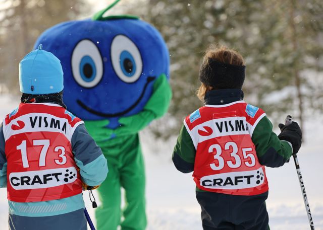 Two children wearing bibs participate in a ski event and are greeted by Orsa Grönklitt’s blueberry mascot Bärra in the snow.