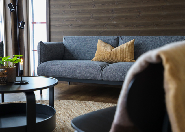 Ett stilrent vardagsrum med grå soffa och en senapsgul kudde. I förgrunden syns ett runt soffbord och en fåtölj. Väggarna är klädda i mörkt trä och ljuset strömmar in från fönstret.