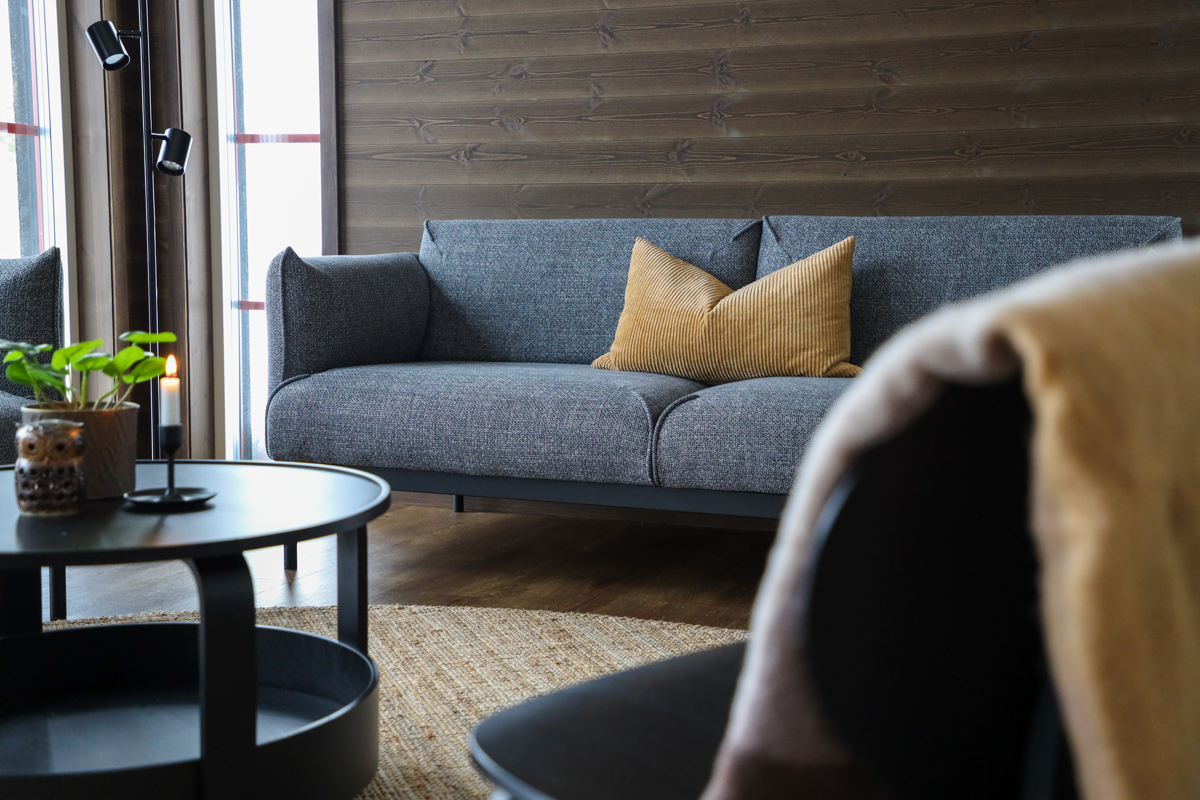 En stilren stue med en grå sofa og en sennepsgul pude. I forgrunden ses et rundt sofabord og en lænestol. Væggene er beklædt med mørkt træ, og dagslys strømmer ind fra vinduet.