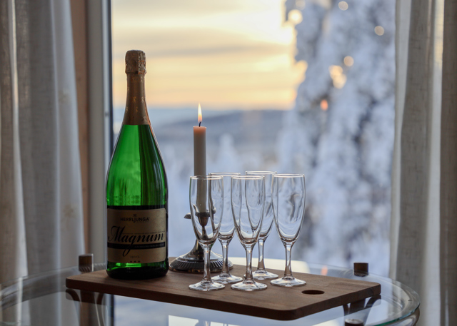 A bottle of champagne, five flutes, and a lit candle on a tray table by a window overlooking a snowy winter landscape.