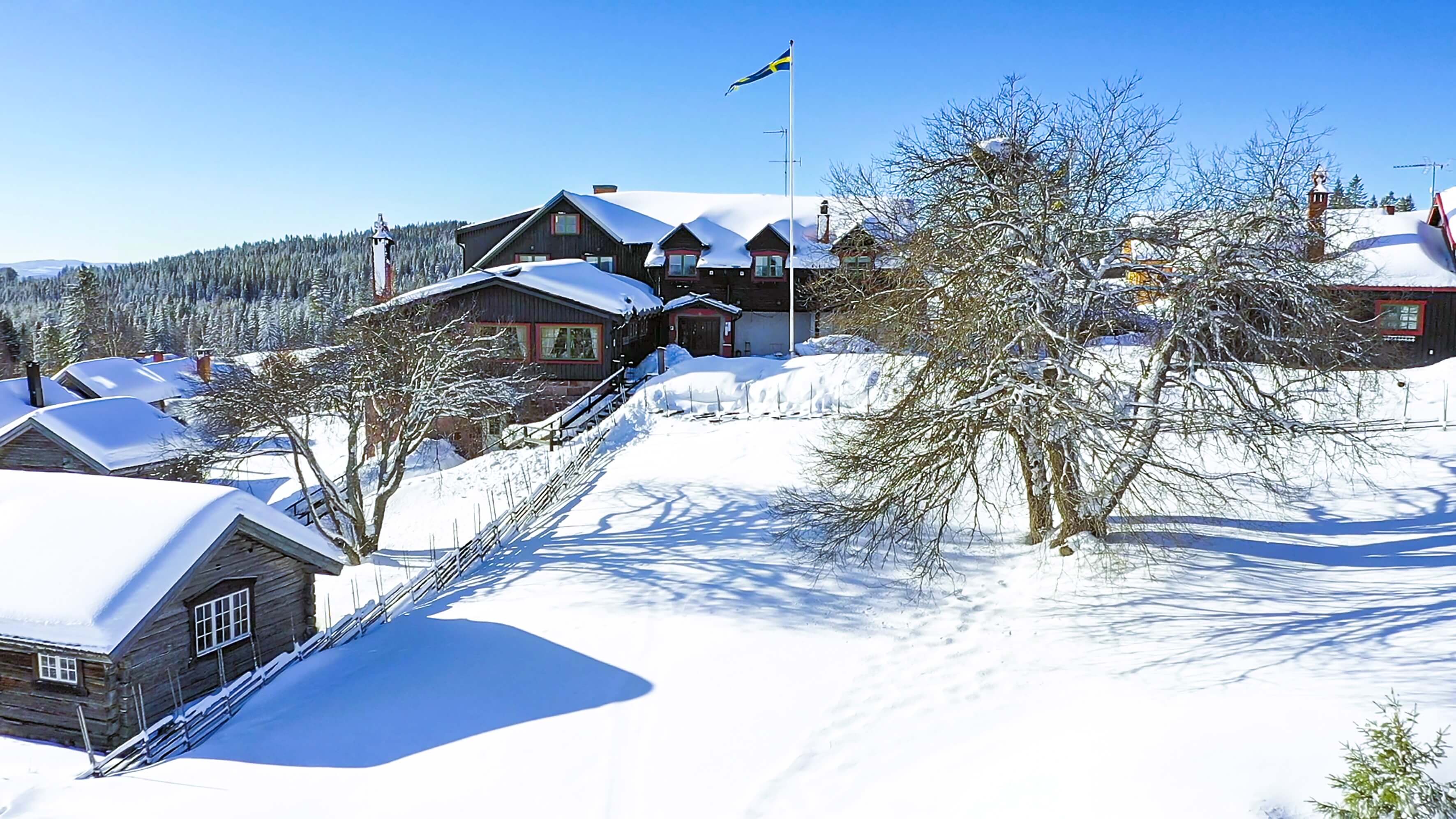 Winter view of Fryksås Hotel & Inn with snow-covered wooden houses and surrounding forest in sunlight.