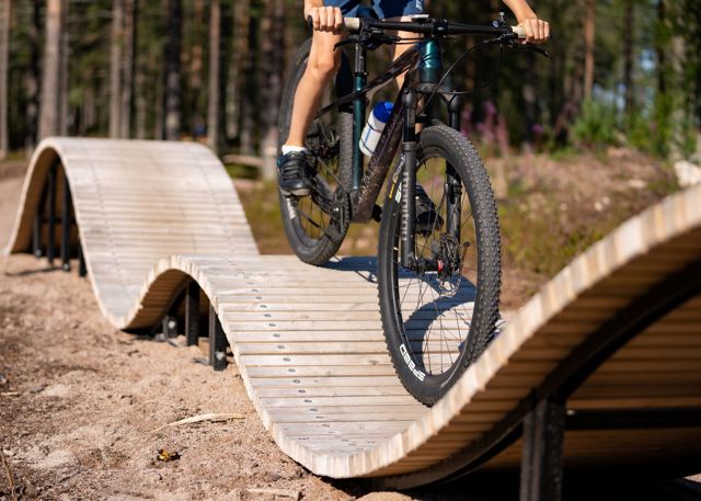 Mountainbikecyklist kör på en vågformad träbro i en skogsmiljö.