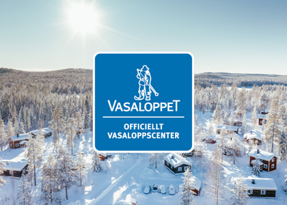 Logotyp för Vasaloppet centrerad över en solig vinterbild med snötäckta stugor och skog i bakgrunden.