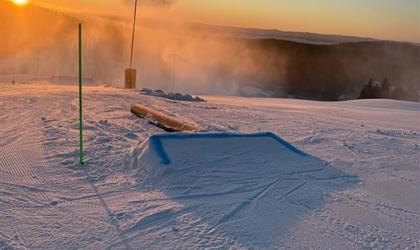 Snötäckt skidområde med ett mindre hopp, snökanon och låg sol vid horisonten.