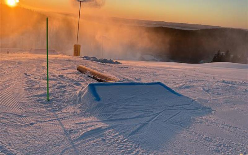 Snötäckt skidområde med ett mindre hopp, snökanon och låg sol vid horisonten.