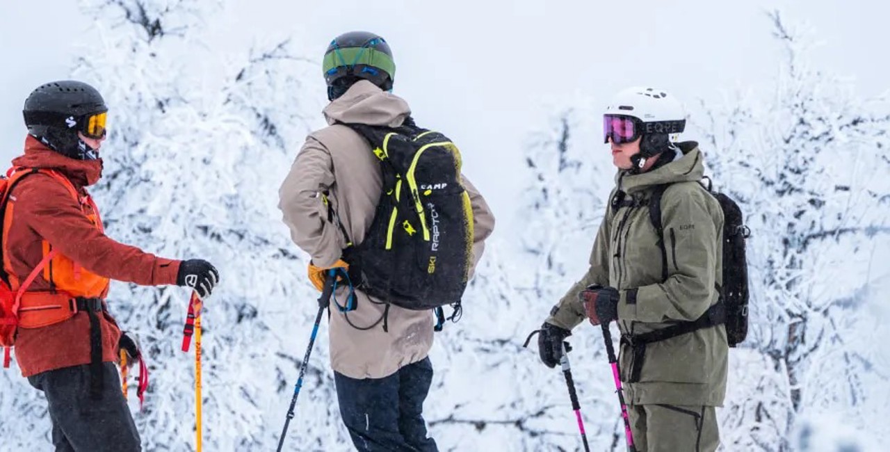 Tre skidåkare med hjälmar, stavar och ryggsäckar i ett snötäckt vinterlandskap.