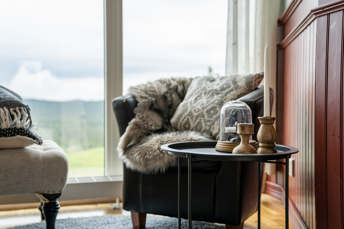 Fåtölj med kudde och fäll bredvid ett bord med ljusstakar vid stort fönster.