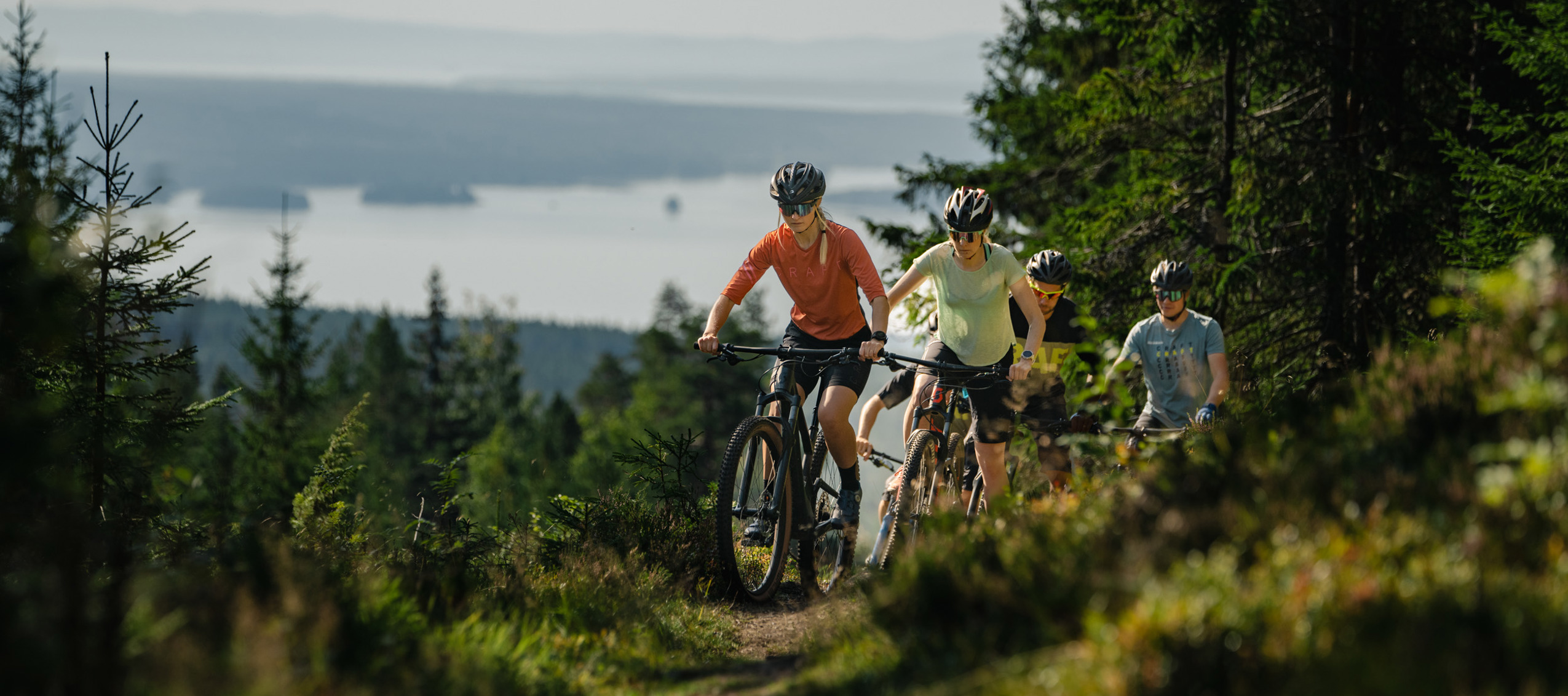Cyklande familj på skogsstig med utsikt över sjö och skogsklädda höjder i Orsa Grönklitt.
