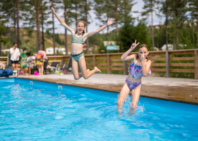 To børn i badetøj hopper glade ned i en udendørs pool på en solrig dag.