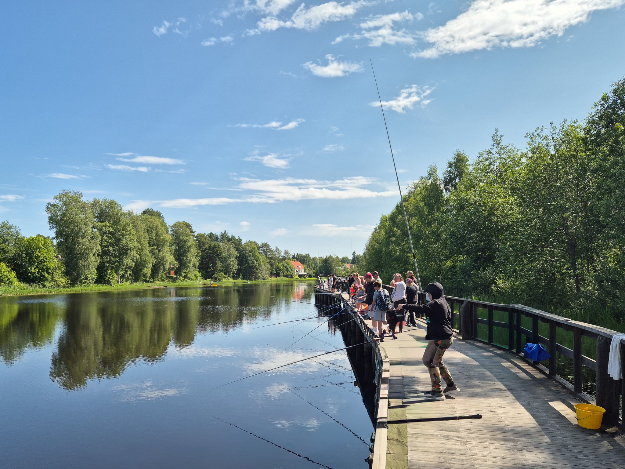 Personer fiskar från en brygga vid en lugn sjö med träd längs stranden.