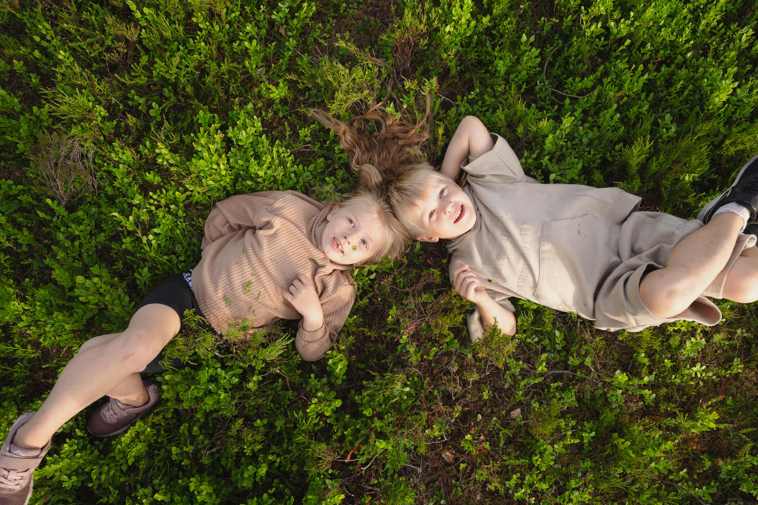 Två barn ligger avslappnat på rygg i tät grönska och tittar upp mot himlen.