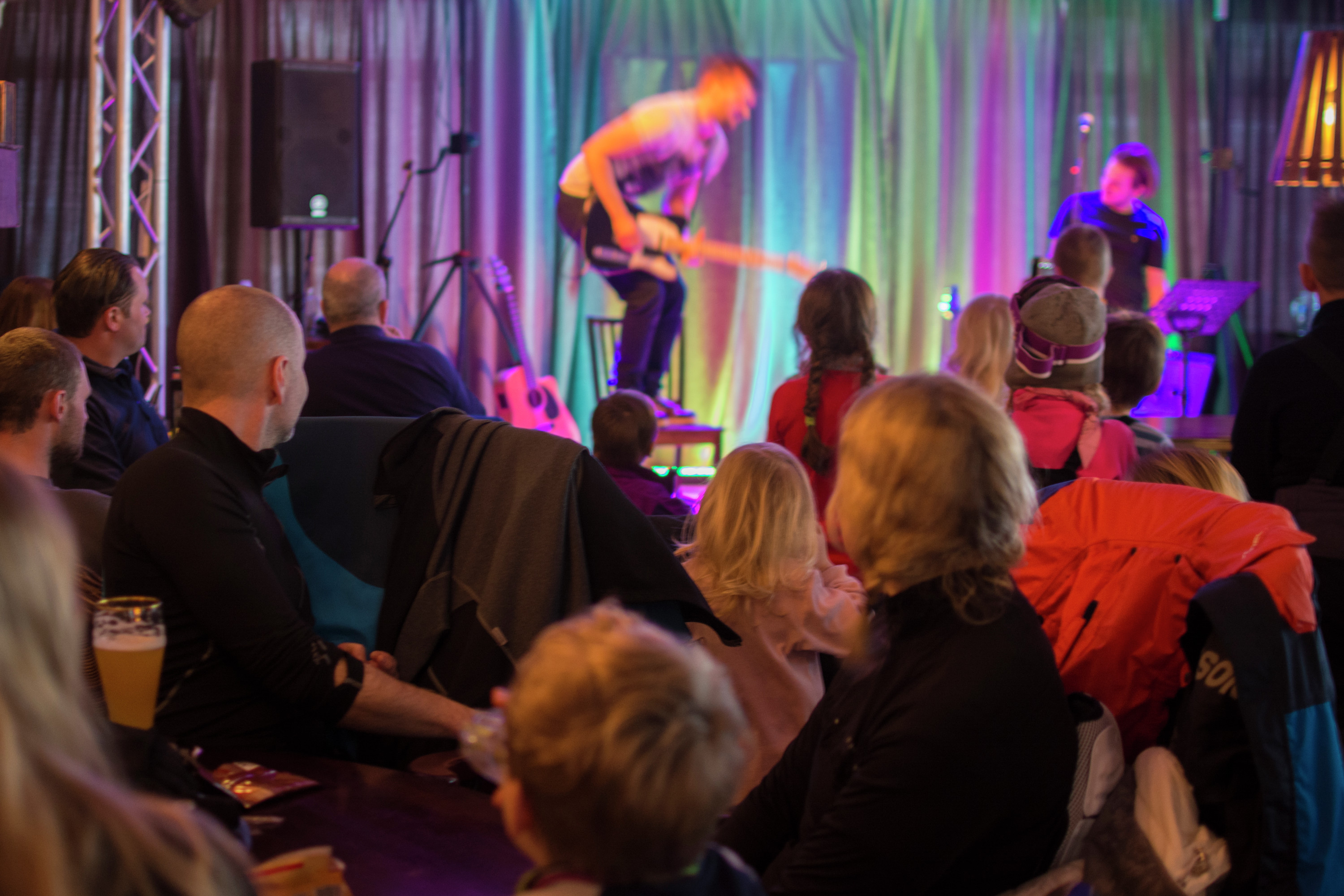 Publik med både vuxna och barn sitter och tittar på en färgglad scenshow med livemusik.