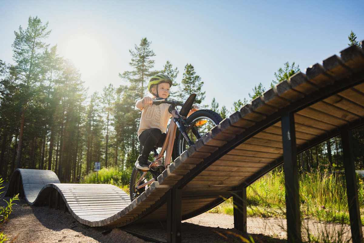 Barn med cykelhjälm cyklar uppför en svängd träbro i soligt väder, omgiven av grönska och tallar.