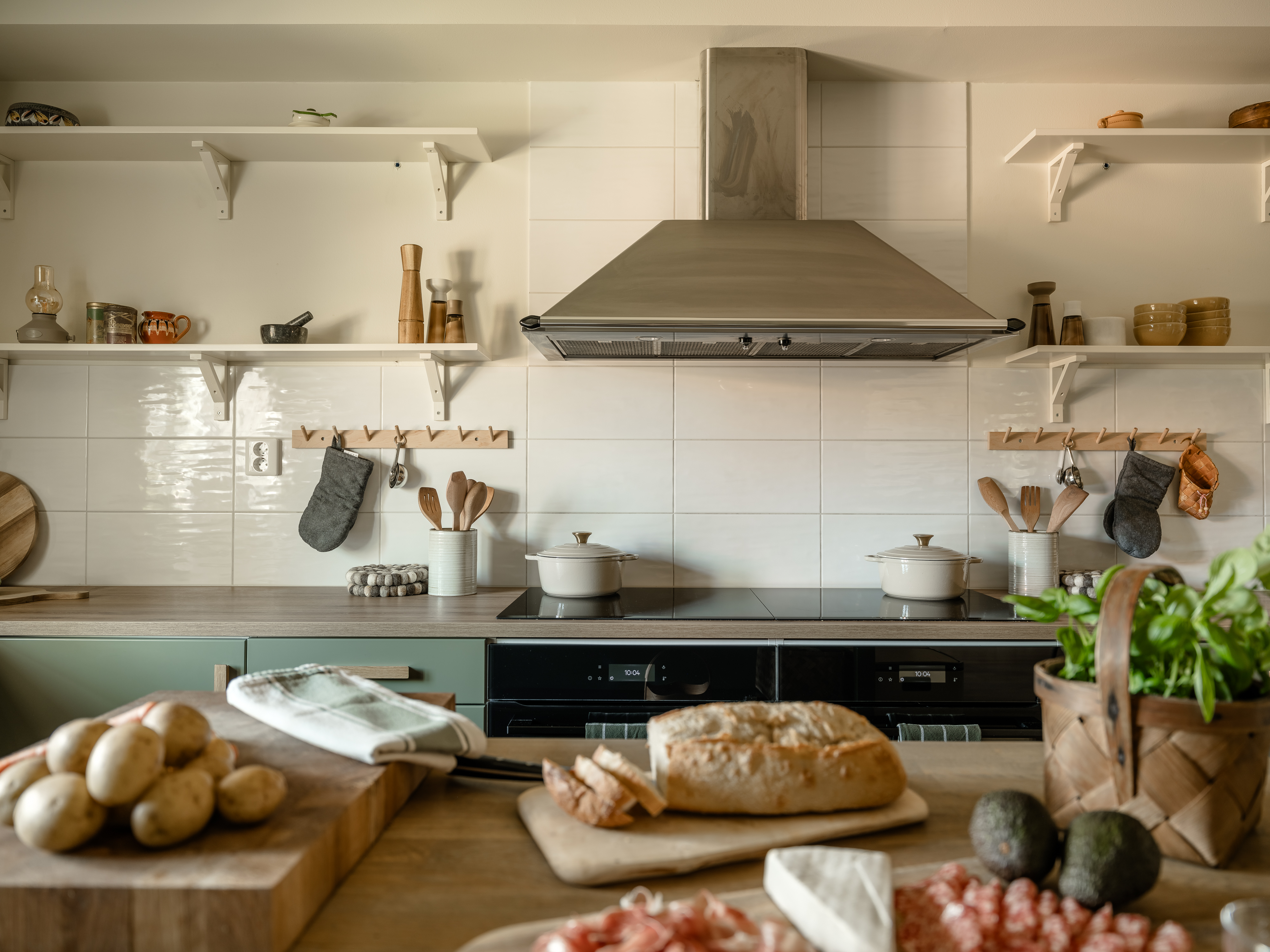 Kök med vita kakelväggar, öppna hyllor och en stor köksfläkt, med matlagningsingredienser som bröd, potatis, basilika och charkuterier framdukade på arbetsbänken.