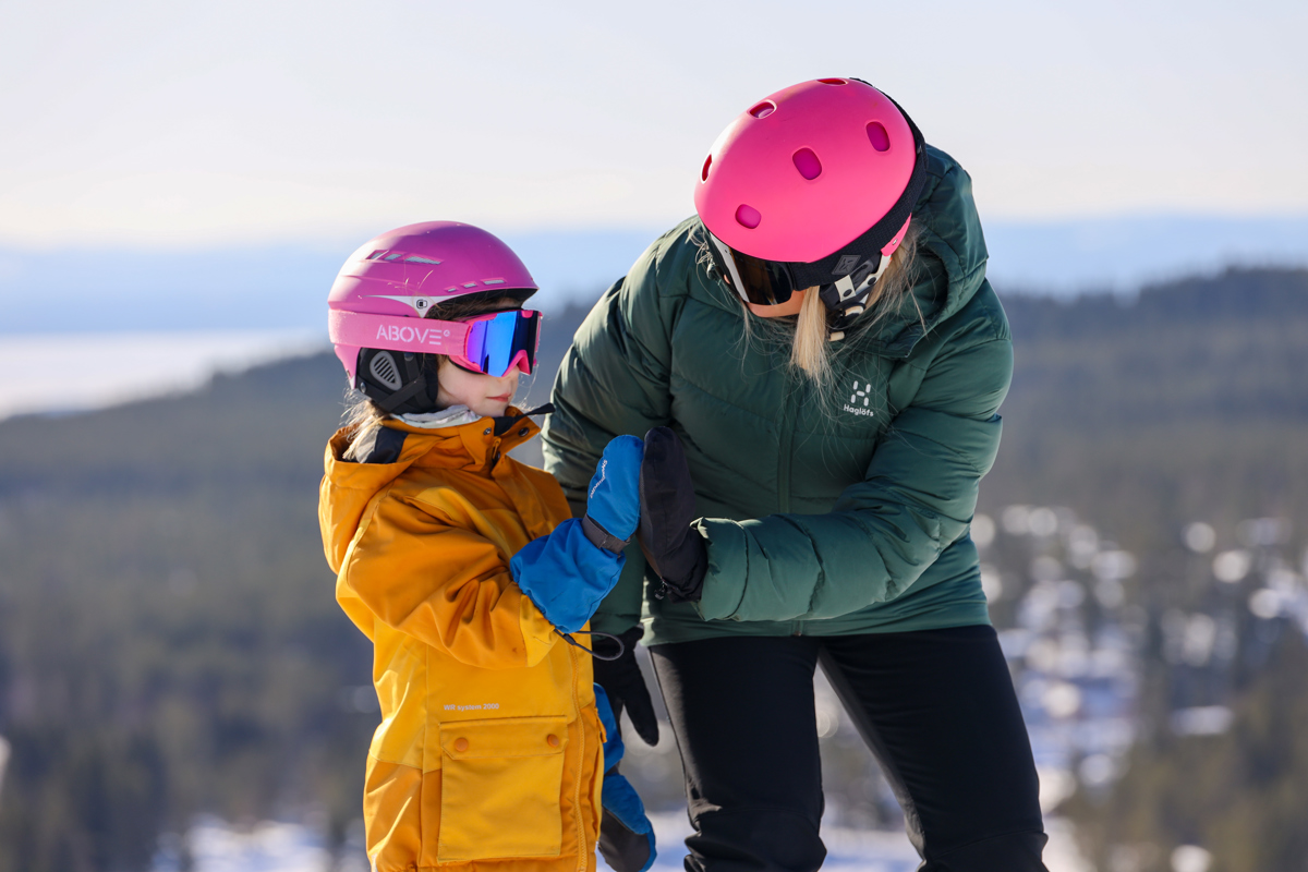 Kvinde og barn med lyserøde hjelme giver high five på skipisten, med snedækket landskab og udsigt i baggrunden.