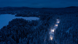 Upplyst väg genom snötäckt skog i skymning, sedd från luften, med fordon som kör på vägen.