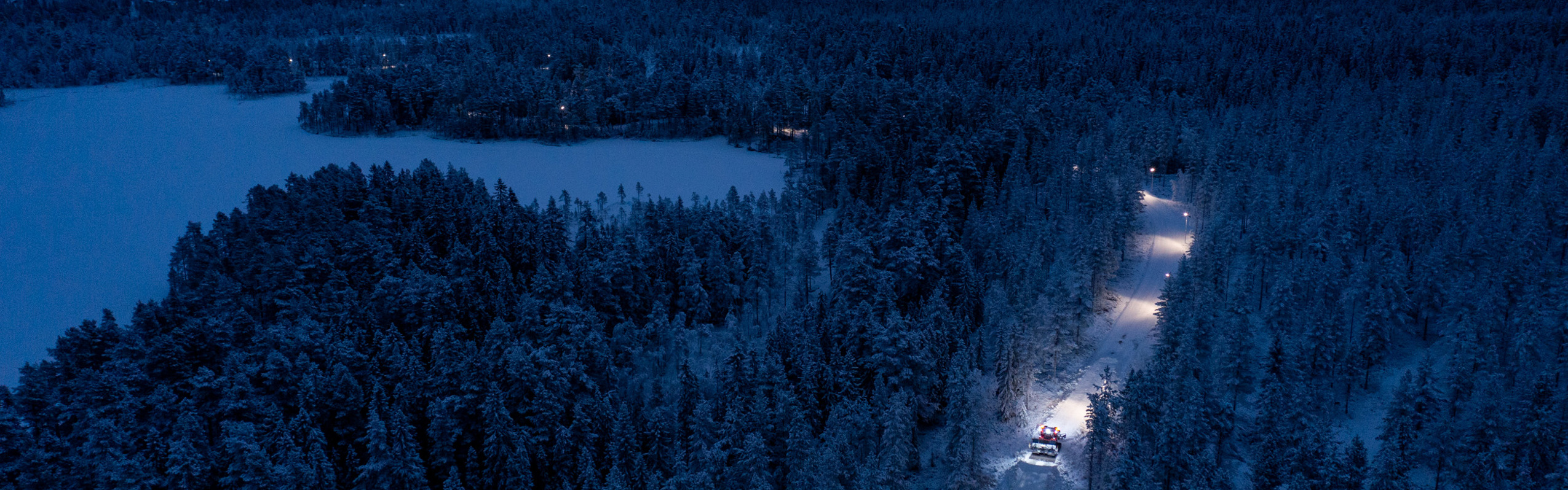 Upplyst väg genom snötäckt skog i skymning, sedd från luften, med fordon som kör på vägen.