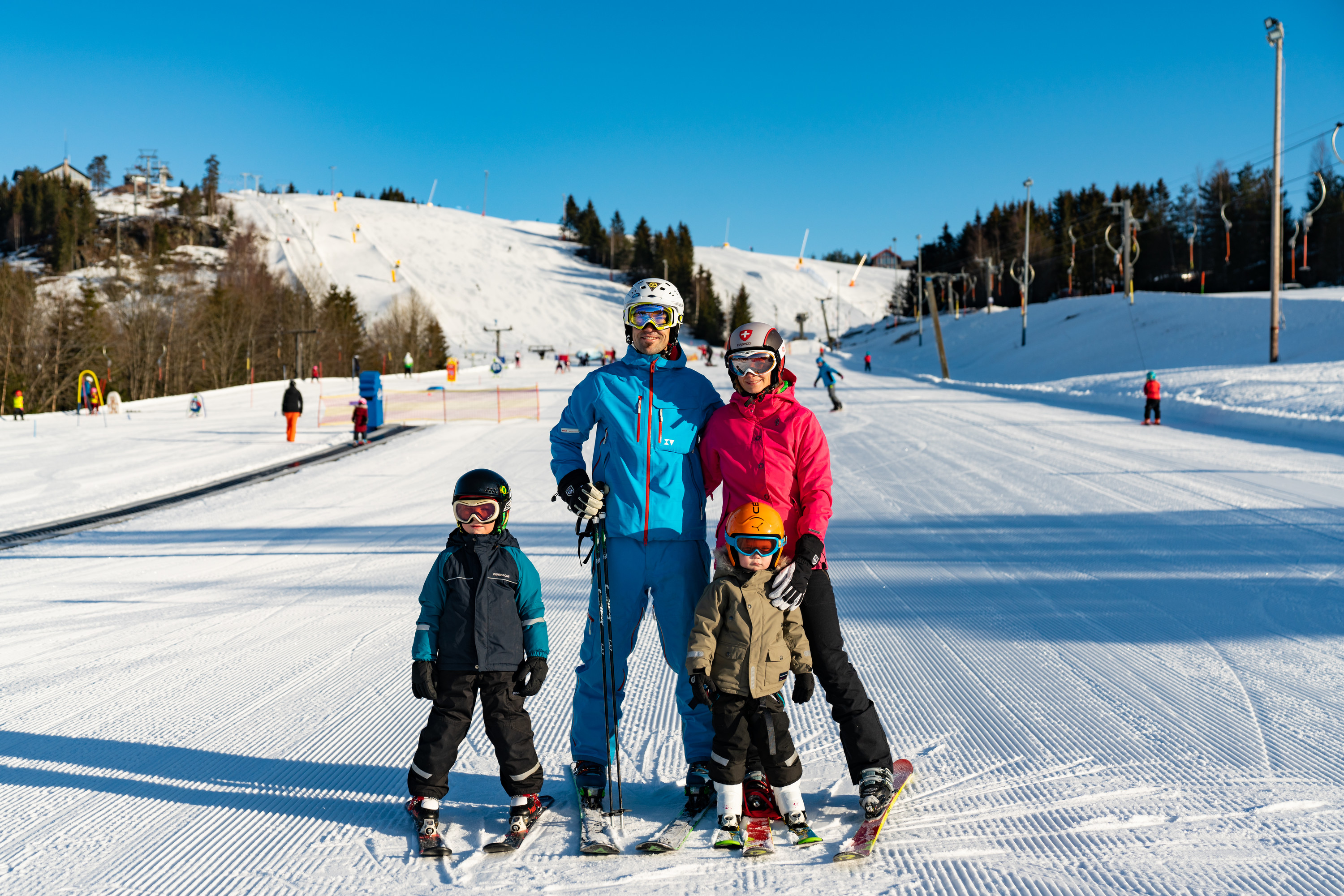 A family with two children stands on skis in a sunny ski slope with groomed runs in the background.