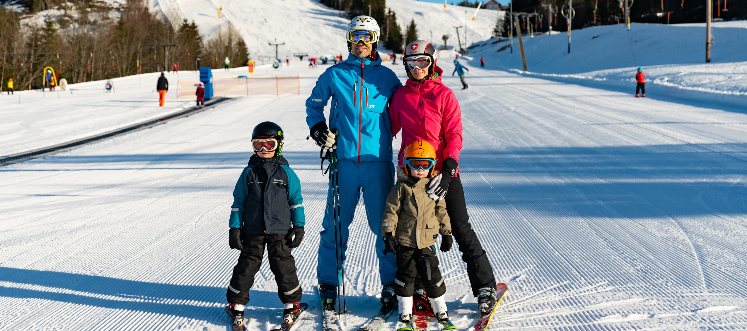 En familj med två barn står på skidor i en solig skidbacke med preparerade pister i bakgrunden.