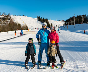 En familj med två barn står på skidor i en solig skidbacke med preparerade pister i bakgrunden.