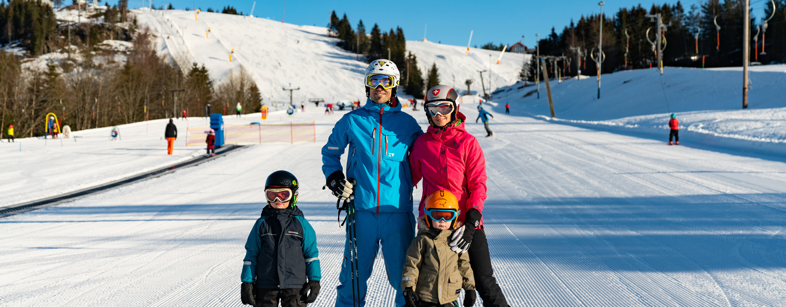 A family with two children stands on skis in a sunny ski slope with groomed runs in the background.