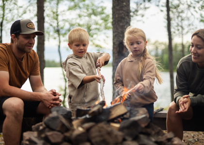 En familj sitter runt en lägereld vid en sjö, där två barn grillar pinnbröd medan de vuxna tittar på och ler.