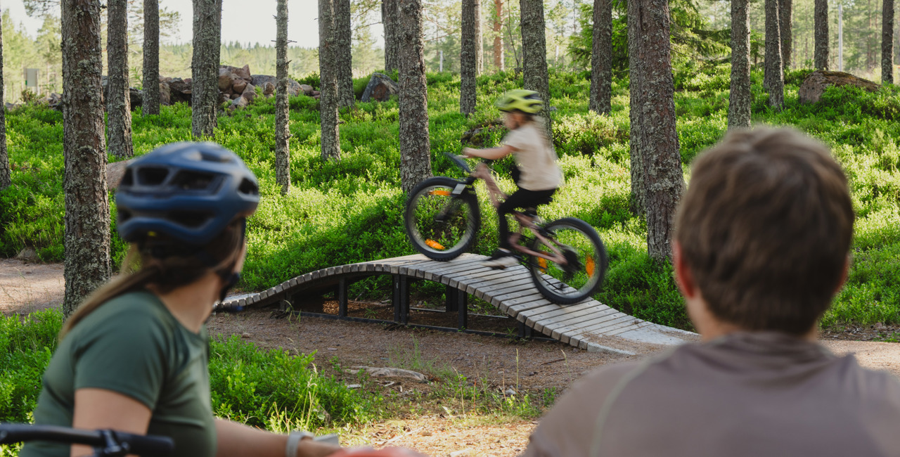 Två vuxna med cykelhjälmar tittar på ett barn som cyklar över en smal träbro i en grönskande skog.