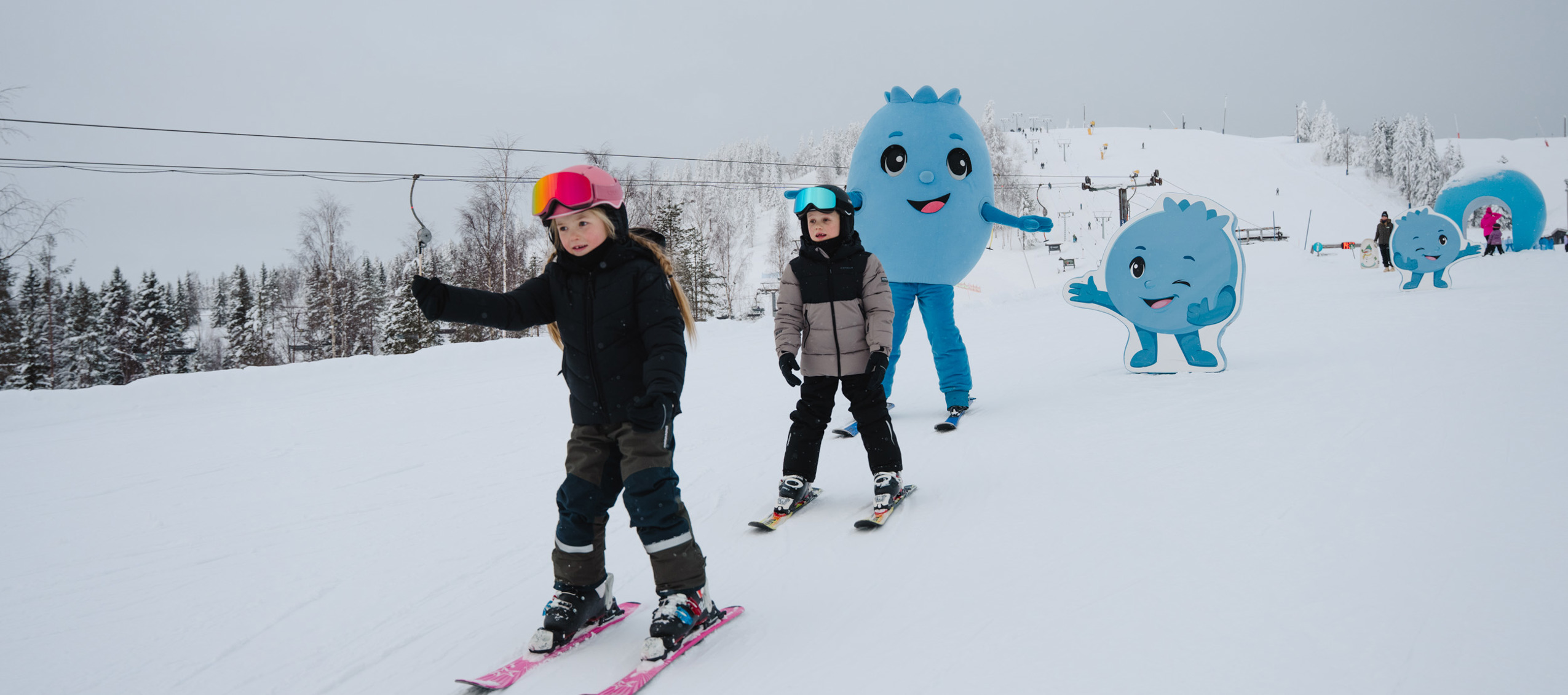 Børn står på ski i en snedækket bakke sammen med en blå figur i baggrunden.