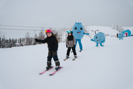 Barn åker skidor i en snötäckt backe tillsammans med en blå figur i bakgrunden.