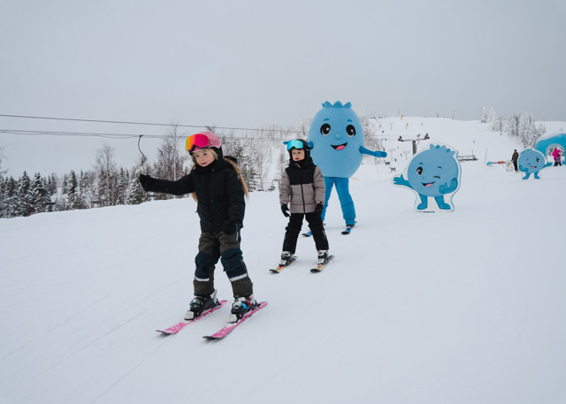 Barn åker skidor i en snötäckt backe tillsammans med en blå figur i bakgrunden.