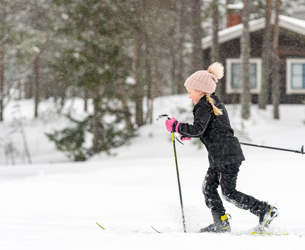 ”Barn i svart skidkläder och rosa vantar åker längdskidor i snöfall framför en timmerstuga.”
