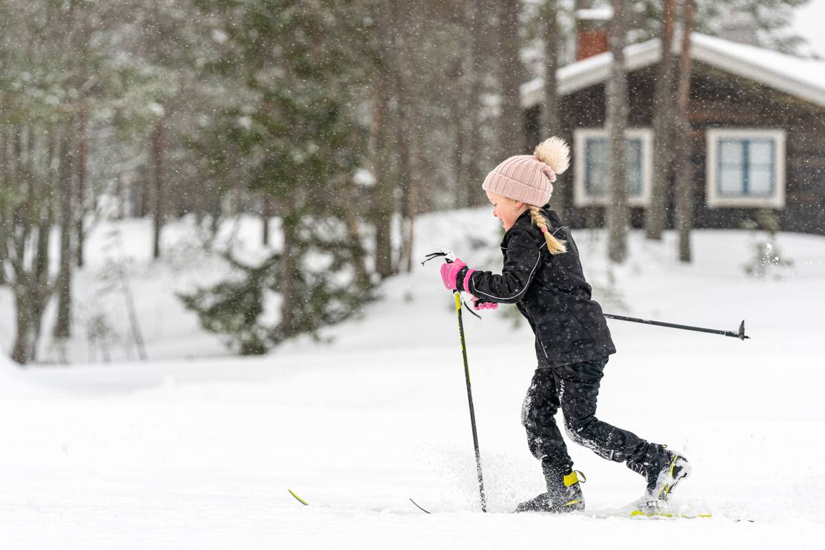 ”Barn i svart skidkläder och rosa vantar åker längdskidor i snöfall framför en timmerstuga.”