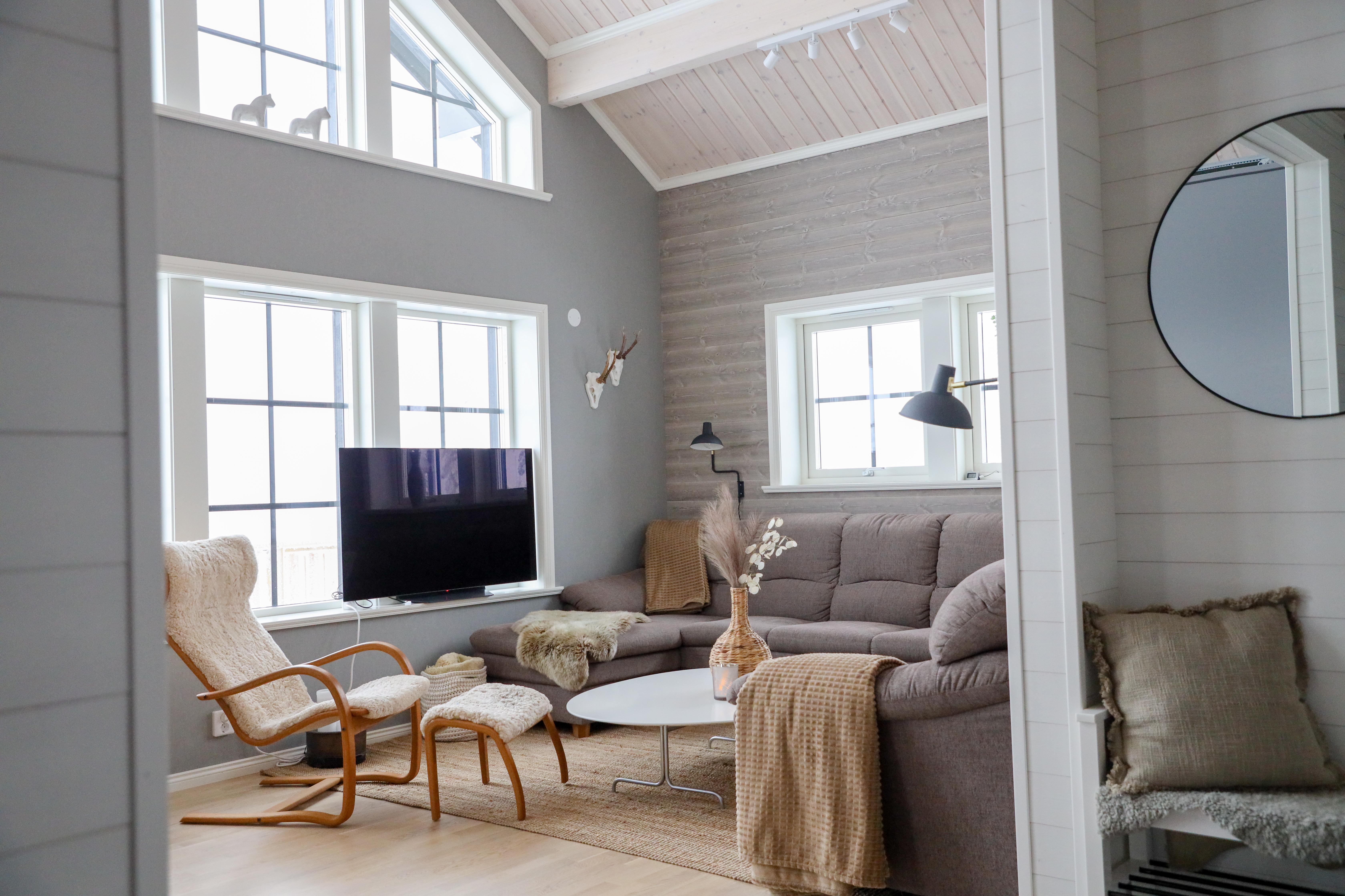 Ljust vardagsrum med hörnsoffa, fåtölj, TV och stora fönster.